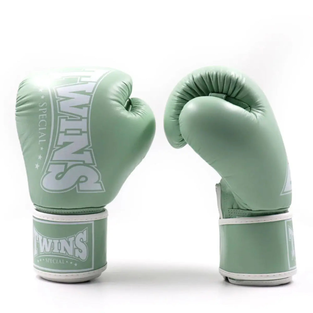 Twee Twins bokshandschoenen in mintgroene kleur, rechtopstaand op een witte achtergrond. De ene handschoen toont het Twins-logo aan de voorkant, terwijl de andere de gebogen pasvorm en polssluiting laat zien, ontworpen voor comfort en duurzaamheid tijdens bokstrainingen.