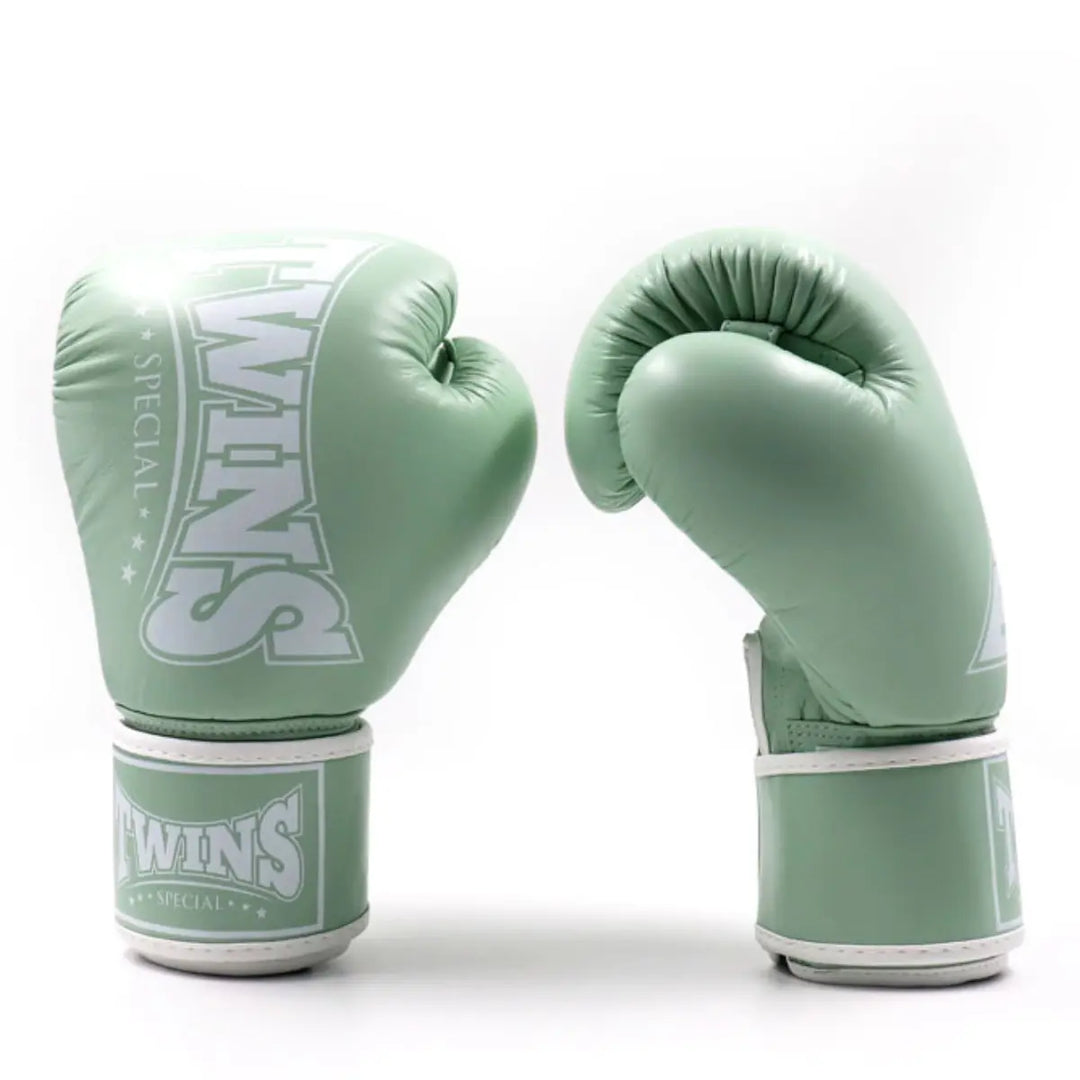 Twee Twins bokshandschoenen in mintgroene kleur, rechtopstaand op een witte achtergrond. De ene handschoen toont het Twins-logo aan de voorkant, terwijl de andere de gebogen pasvorm en polssluiting laat zien, ontworpen voor comfort en duurzaamheid tijdens bokstrainingen.