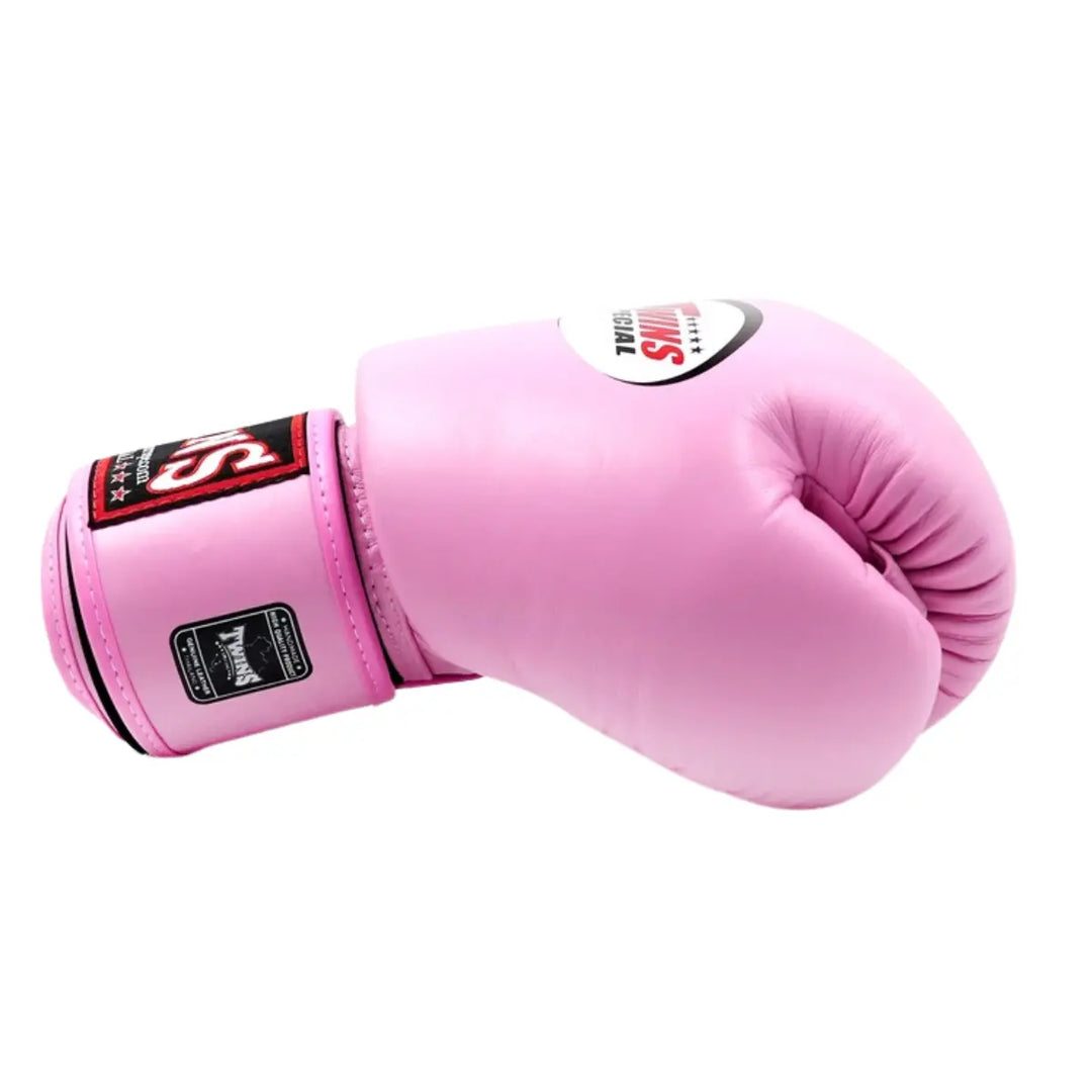De Twins Bokshandschoen Roze BGVL3 ligt zijwaarts op een witte achtergrond, met het Twins Special-logo duidelijk zichtbaar op de bovenkant. De handschoen heeft een gladde roze leren afwerking en een stevige polssluiting met zwart-rode accenten. Deze foto benadrukt de kwaliteit, duurzaamheid en vrouwelijke uitstraling van de professionele bokshandschoen.