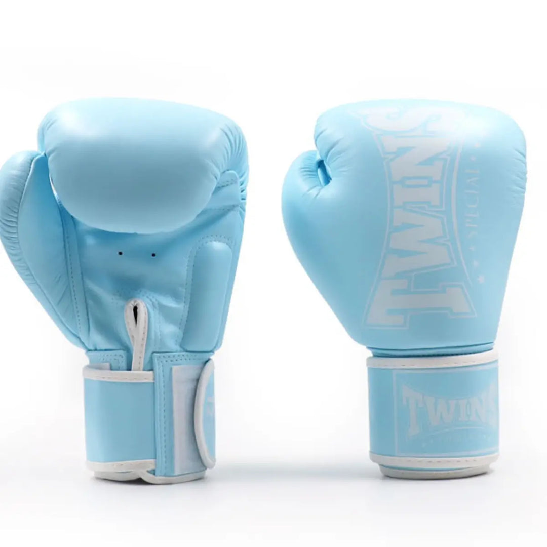 Twee Twins bokshandschoenen in babyblauw met witte accenten, weergegeven van voor- en achteraanzicht. Het Twins-logo is subtiel zichtbaar op de polsband, terwijl de binnenzijde ademende ventilatiegaatjes en stevige polssteun toont voor optimale prestaties tijdens bokstraining.
