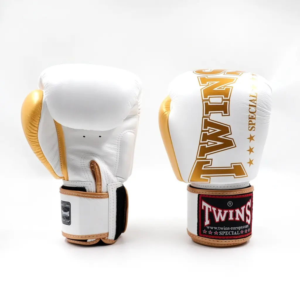 Twee Twins bokshandschoenen in wit en goud, rechtopstaand op een witte achtergrond met groot gouden TWINS-logo op de vuistzijde en rood label op de polsband.