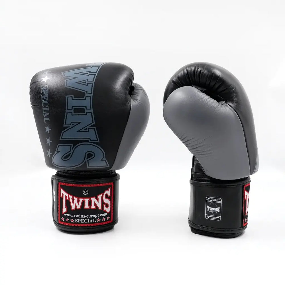 Set zwart-grijze Twins bokshandschoenen, rechtopstaand op een witte achtergrond, met groot TWINS-logo op de hand en rood label op de polsband.