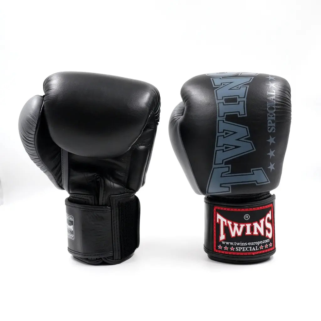 Twee zwart-grijze Twins bokshandschoenen op witte achtergrond, met rood Twins-logo op de polsband en groot TWINS-opdruk op de vuistzijde.