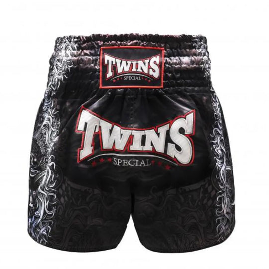 Achteraanzicht van het Twins Special kickboksbroekje in zwart, afgewerkt met een subtiele drakenprint aan de zijkanten en een extra groot TWINS-logo in rood-wit op de achterkant. De brede witte elastische tailleband met rood TWINS-label zorgt voor een stevige pasvorm. Gemaakt van hoogwaardig satijn polyester voor lichtheid en flexibiliteit tijdens elke training. Een statement short voor de kickbokser die kracht wil uitstralen.