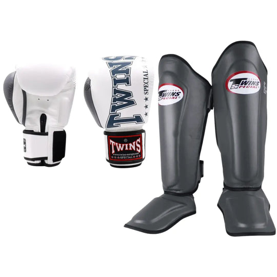 Set van Twins Special bokshandschoenen in wit met zwarte accenten en grijze scheenbeschermers met rood-wit logo, geschikt voor kickboksen en MMA.