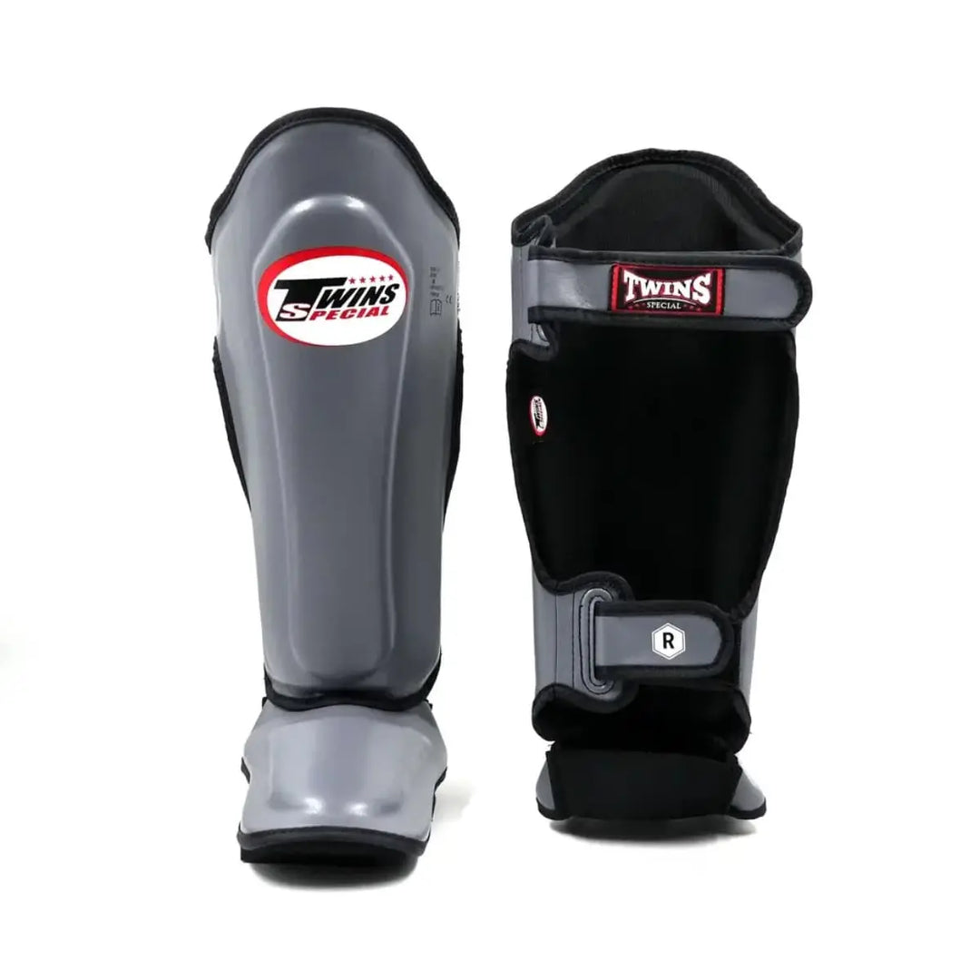 Twins Special SGL7 scheenbeschermers in grijs, getoond van voor- en achterzijde met zwart gevoerde binnenkant. Voorzien van stevige padding, geïntegreerde voetbescherming en dubbele klittenbandsluiting voor een stabiele pasvorm tijdens kickbokstraining en sparren.
