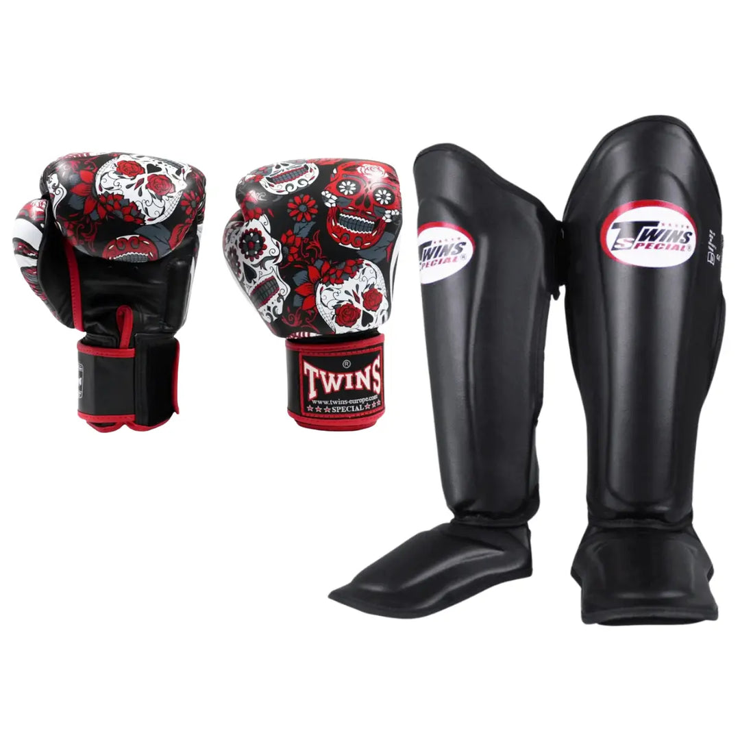 Productfoto van een Twins kickboks set met bokshandschoenen voorzien van rood, wit en zwarte skull print en zwarte scheenbeschermers met Twins Special logo, afgebeeld tegen een witte achtergrond.