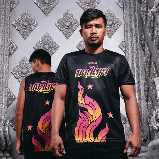 Afbeelding van twee modellen die het Yokkao The Flame workout T-shirt in zwart dragen, getoond in voor- en achteraanzicht. Het shirt heeft een opvallende vlamprint in roze en geel met Thaise tekst en sterdetails. De sportieve pasvorm en het krachtige design zorgen voor een dynamische uitstraling tijdens trainingen en fotoshoots.