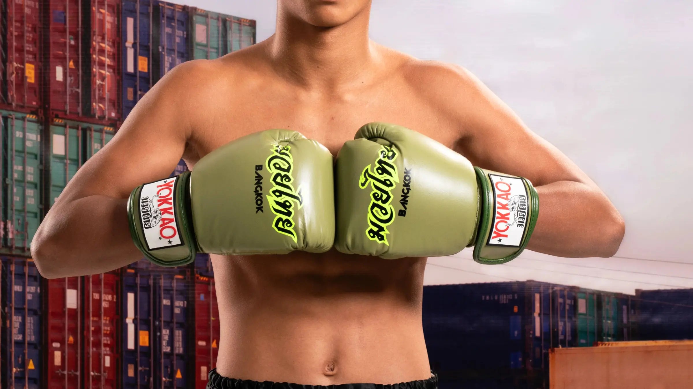 Deze afbeelding toont een sporter met de armen naar voren gebracht, waarbij een paar olijfgroene YOKKAO bokshandschoenen centraal in beeld staat. De handschoenen hebben Thaise lettering en opvallende branding op de polsband. Op de achtergrond zijn gestapelde zeecontainers zichtbaar, wat de foto een stoere, industriële sfeer geeft.