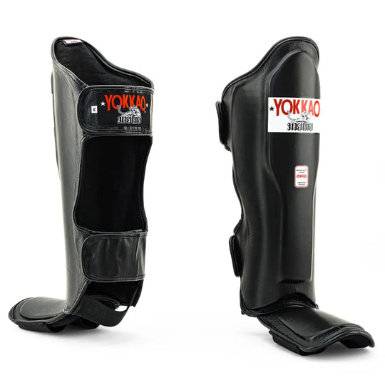 A pair of professional black leather shin guards for Muay Thai and kickboxing. The guards feature a contoured design for protection, secure hook-and-loop straps at the back, and the red and white YOKKAO brand logo on the front.