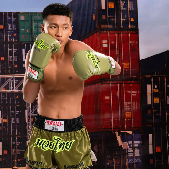 Kickbokser met Yokkao Bangkok Groen Bokshandschoenen en bijpassende short in gevechtshouding. De legergroene uitrusting met neonaccenten toont het kenmerkende Yokkao-design, perfect voor intensieve bokstrainingen en professionele sparringsessies.