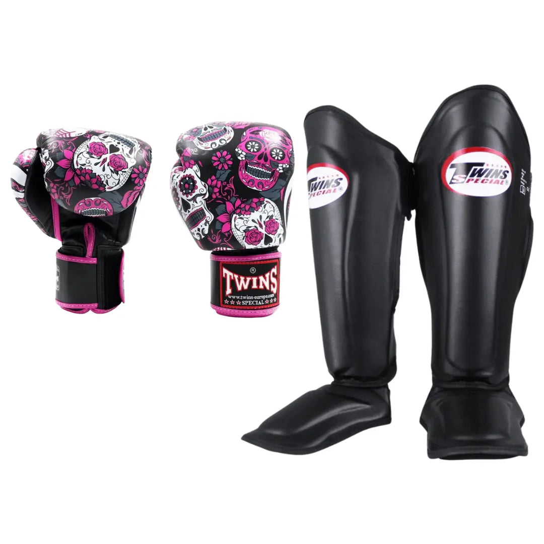 Productfoto van een Twins kickboks set bestaande uit bokshandschoenen met roze, wit en zwarte skull print en zwarte scheenbeschermers met Twins Special logo, weergegeven tegen een witte achtergrond.