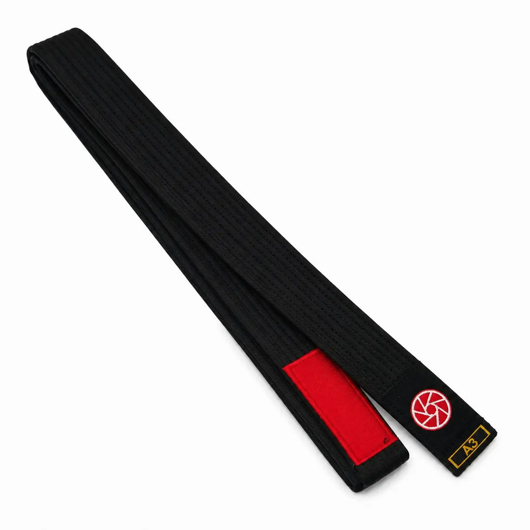 Zwarte BJJ band in maat A3 met rood vlak voor graduatiestrepen en een rood-wit logo op het label. De lange, stevige geweven structuur maakt de band geschikt voor BJJ-trainingen, graduaties en dagelijks gebruik op de mat.