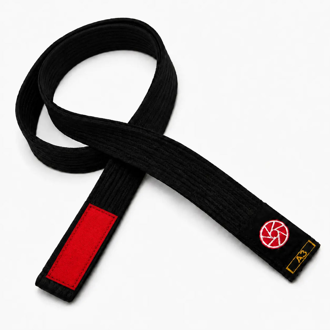 Zwarte BJJ band in maat A3 met rood vlak voor graduatiestrepen en een rood-wit logo op het label. De stevige geweven structuur maakt de band geschikt voor BJJ-trainingen, graduaties en dagelijks gebruik op de mat.