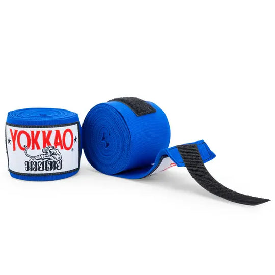 Blauwe Yokkao handwraps gefotografeerd tegen een witte achtergrond, waarbij één bandage is opgerold en de andere deels uitgerold met zichtbare klittenbandsluiting. De witte logo patch met rood YOKKAO-logo en zwarte details is duidelijk zichtbaar. Ideaal voor extra pols- en knokkelondersteuning tijdens boksen en kickboksen.