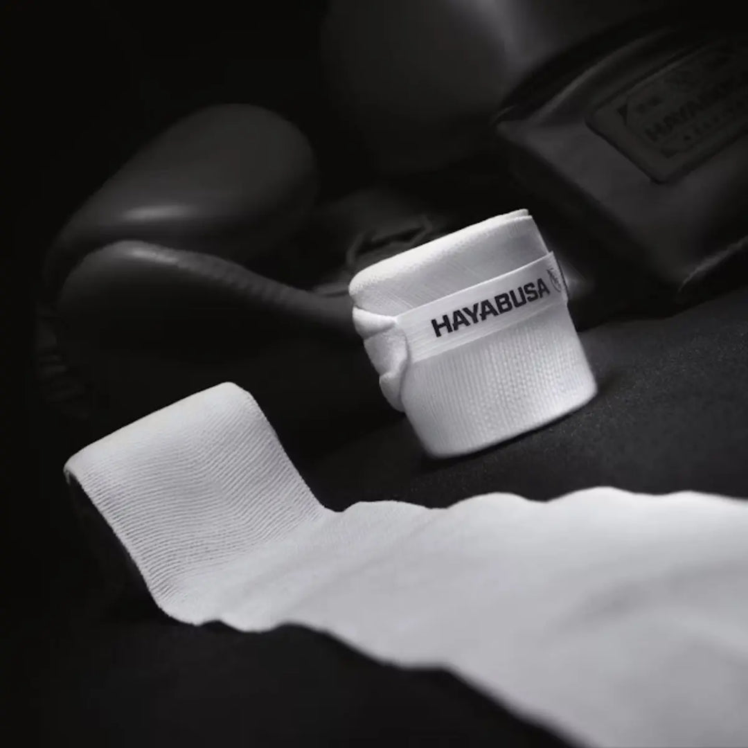 Artistieke, gedempte sfeerfoto van een opgerolde witte Hayabusa handbandage met het zwarte logo, en een onopgerolde band die naar voren wijst. Op de achtergrond is een zwarte bokshandschoen wazig zichtbaar