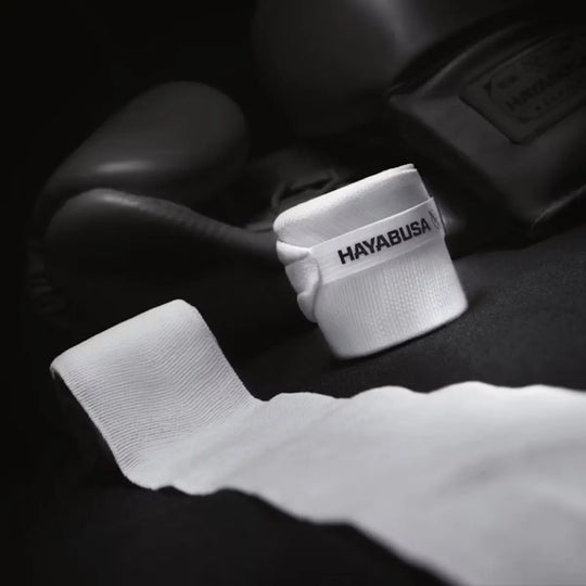 Artistieke, gedempte sfeerfoto van een opgerolde witte Hayabusa handbandage met het zwarte logo, en een onopgerolde band die naar voren wijst. Op de achtergrond is een zwarte bokshandschoen wazig zichtbaar