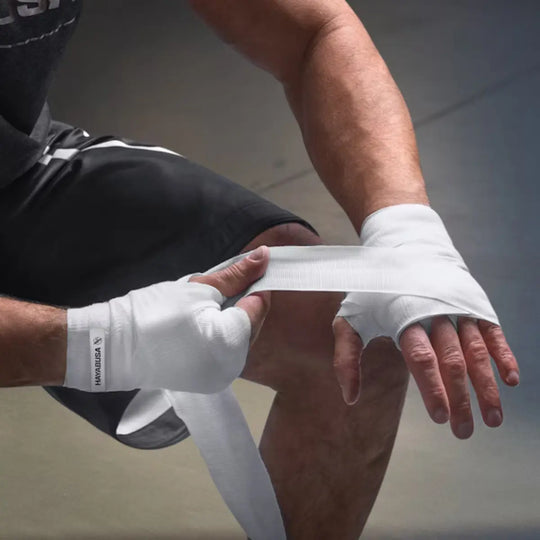 Close-up van een man die zijn hand omwikkelt met witte Hayabusa handwraps, met het Hayabusa-logo op de manchet en de andere handwrap al omgewikkeld