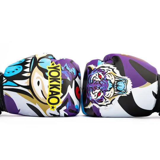 Close-up van twee Yokkao Angry Tiger bokshandschoenen tegen een witte achtergrond. De paarse handschoenen tonen een opvallende tijgerillustratie met rode ogen en blauwe accenten, gecombineerd met het gele YOKKAO-logo. Het grafische design bevat zwart, wit en paarse panelen voor een krachtige uitstraling.