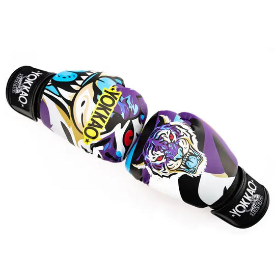 Twee Yokkao Angry Tiger bokshandschoenen diagonaal geplaatst tegen een witte achtergrond. De paarse handschoenen tonen de kleurrijke tijgerprint met rode ogen en het gele YOKKAO-logo op de voorzijde. De zwarte klittenbandsluitingen met merklogo zijn zichtbaar aan beide uiteinden.