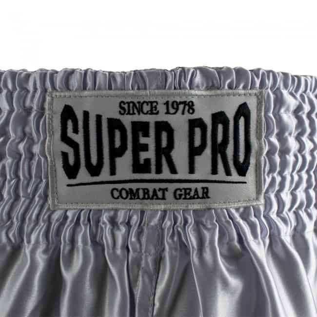 Close-up van het Super Pro Combat Gear logo op de tailleband van het witte Brave kickboksbroekje. Het logo is gestikt op een zilvergrijze achtergrond met zwarte letters. Duidelijk zichtbare elastische band voor stevige pasvorm.