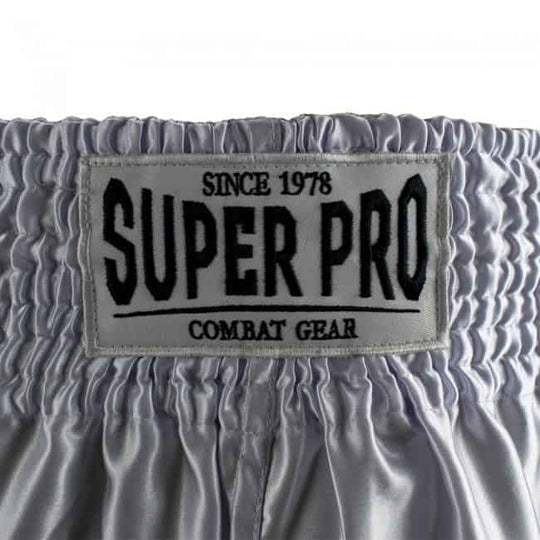 Close-up van het Super Pro Combat Gear logo op de tailleband van het witte Brave kickboksbroekje. Het logo is gestikt op een zilvergrijze achtergrond met zwarte letters. Duidelijk zichtbare elastische band voor stevige pasvorm.