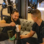 Twee mannen in een sportschool lachen terwijl de een een schepje Whey Water eiwitpoeder van Saevis Nutrition toevoegt aan een shakebeker.
