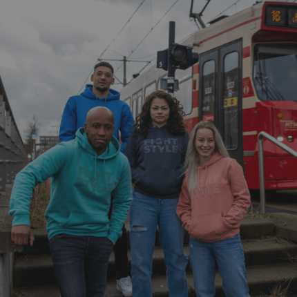 Een groep van vier mensen draagt vrijetijdskleding met de tekst "FIGHT STYLE", poserend voor een tram.