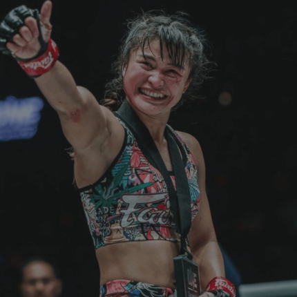 Een vrolijke vrouwelijke vechtsporter viert haar overwinning in de ring met een opgeheven vuist; ze draagt kleurrijke vechtsportkledij en handschoenen, met zichtbare tekenen van een recent gevecht op haar gezicht.