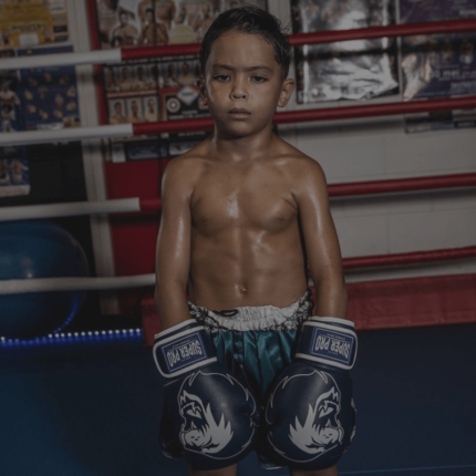 Een jonge jongen met bokshandschoenen staat serieus in een boksring, met posters op de achtergrond.
