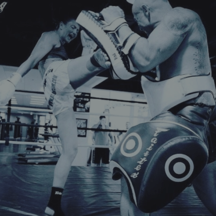 Twee Muay Thai-vechters oefenen in een gymzaal; een vechter levert een hoge trap af op de trapkussens die worden vastgehouden door zijn getatoeëerde partner die beschermende uitrusting draagt.