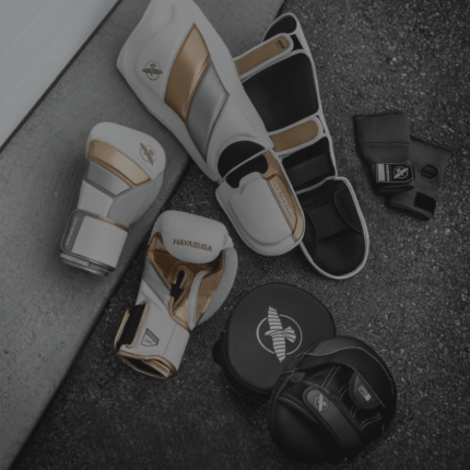 Verschillende vechtsportuitrustingen van Hayabusa op een grijze ondergrond, waaronder wit met gouden scheenbeschermers, zwarte en witte bokshandschoenen, polsbandages en een hoofdbeschermer met het merklogo.