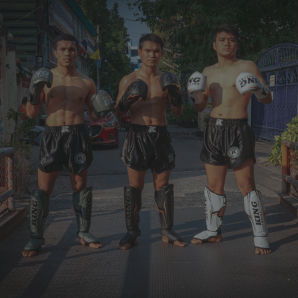 Drie mannen in traditionele Muay Thai-houding, gekleed in zwarte shorts en met bokshandschoenen en scheenbeschermers, staan klaar om te trainen op een straat in Thailand.